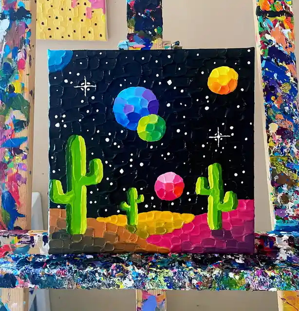 A thickly textured, colorful painting on a canvas depicts a surreal desert scene with green cacti on a multi-hued ground…
