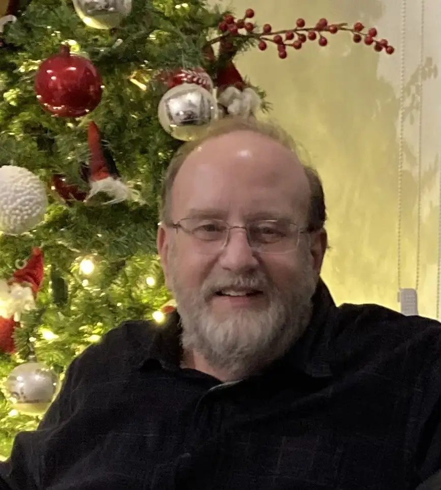 a middle-aged man with glasses and a beard smiles while sitting in front of a decorated christmas tree with red and silver…