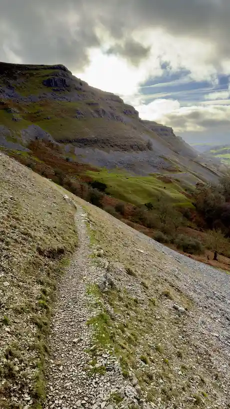 A steep, rocky hiking trail winds up a grassy, rocky mountainside under a cloudy sky with patches of sunlight. The path is…