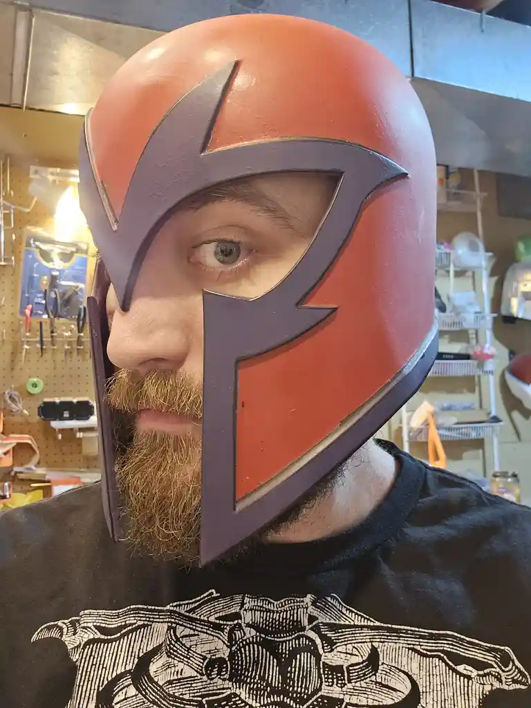 A man with a full beard and green eyes wears a red and purple helmet resembling Magneto's from X-Men, covering his forehead…