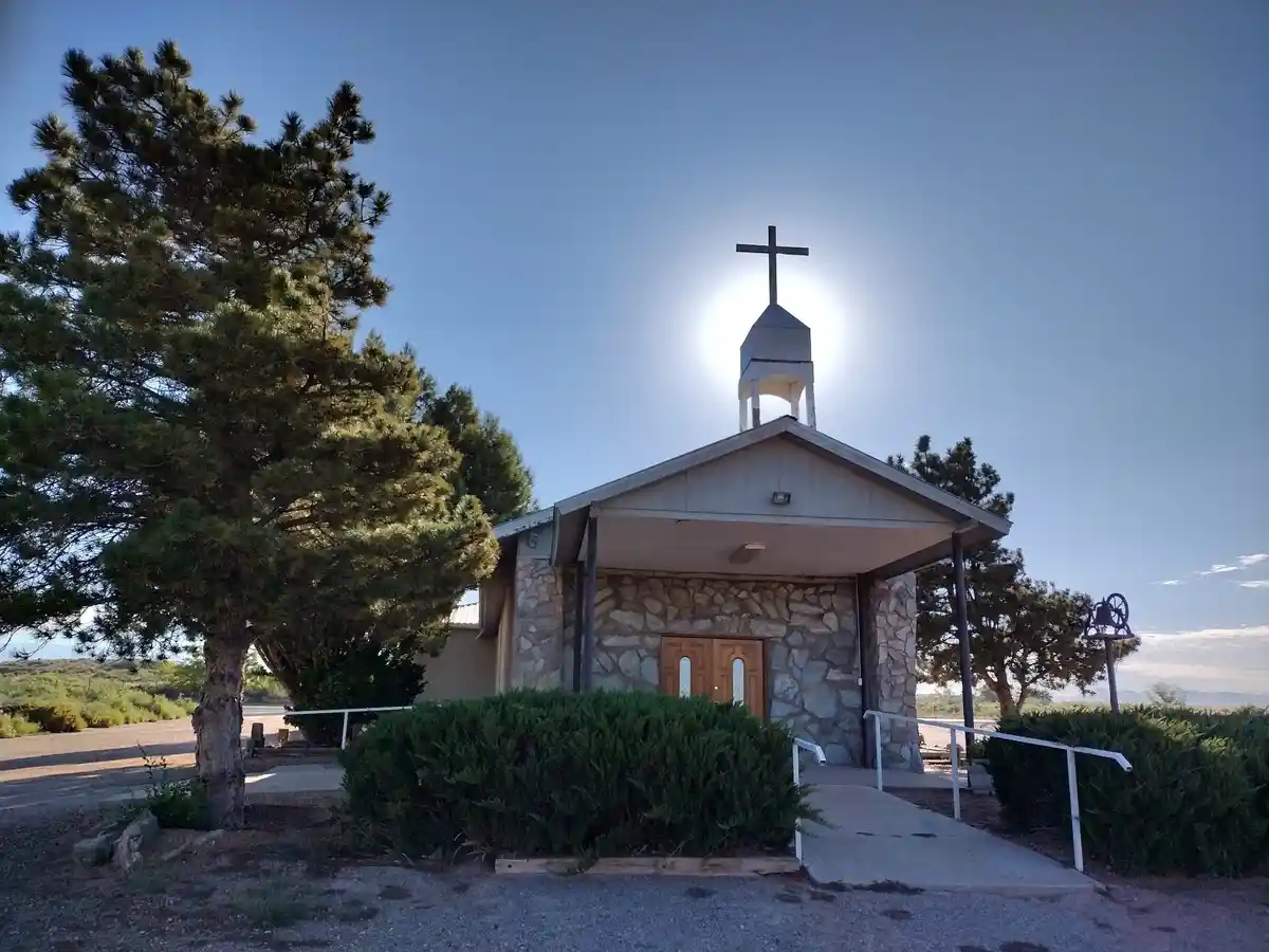 A small stone church with a wooden door and a cross atop its steeple is pictured under a clear blue sky. The sun is…