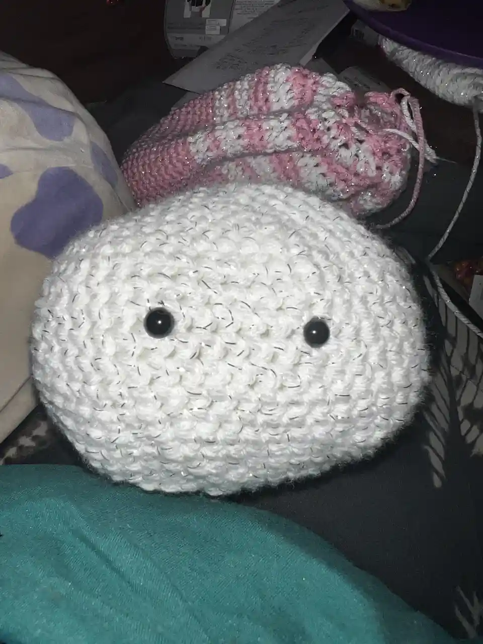A white, round, crocheted plushie with two black button eyes sits on a surface, looking like a simple, slightly lopsided…