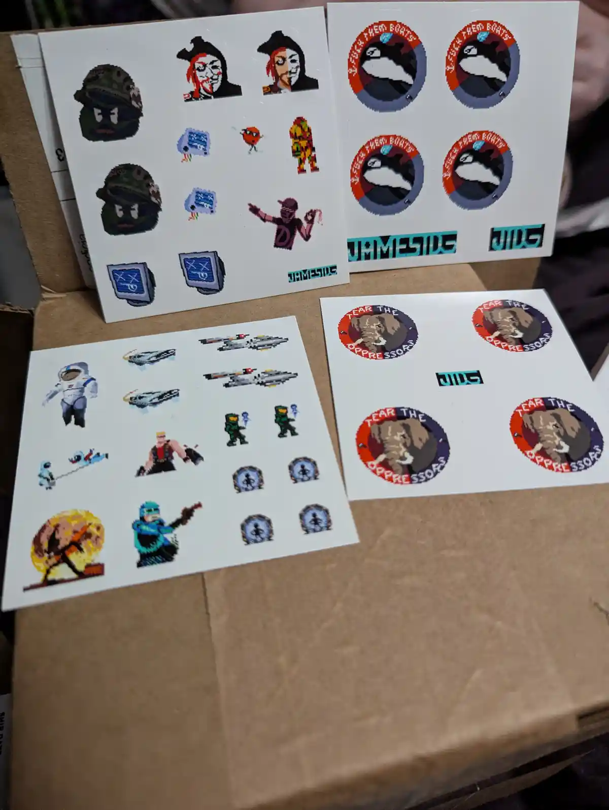 Four sheets of pixel art stickers are displayed on a cardboard box. The sheets feature various retro video game characters…