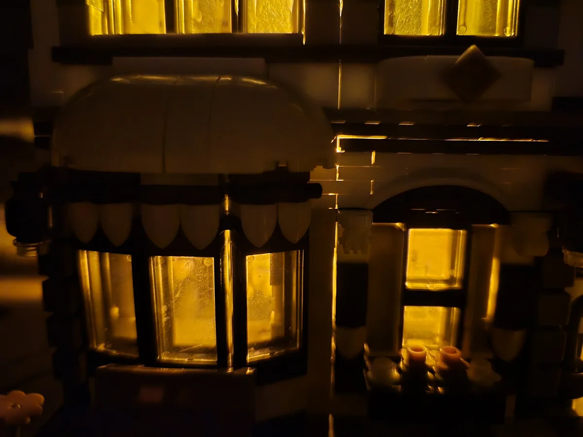 A close-up of a detailed LEGO house model at night, illuminated from within by warm yellow lights visible through multiple…