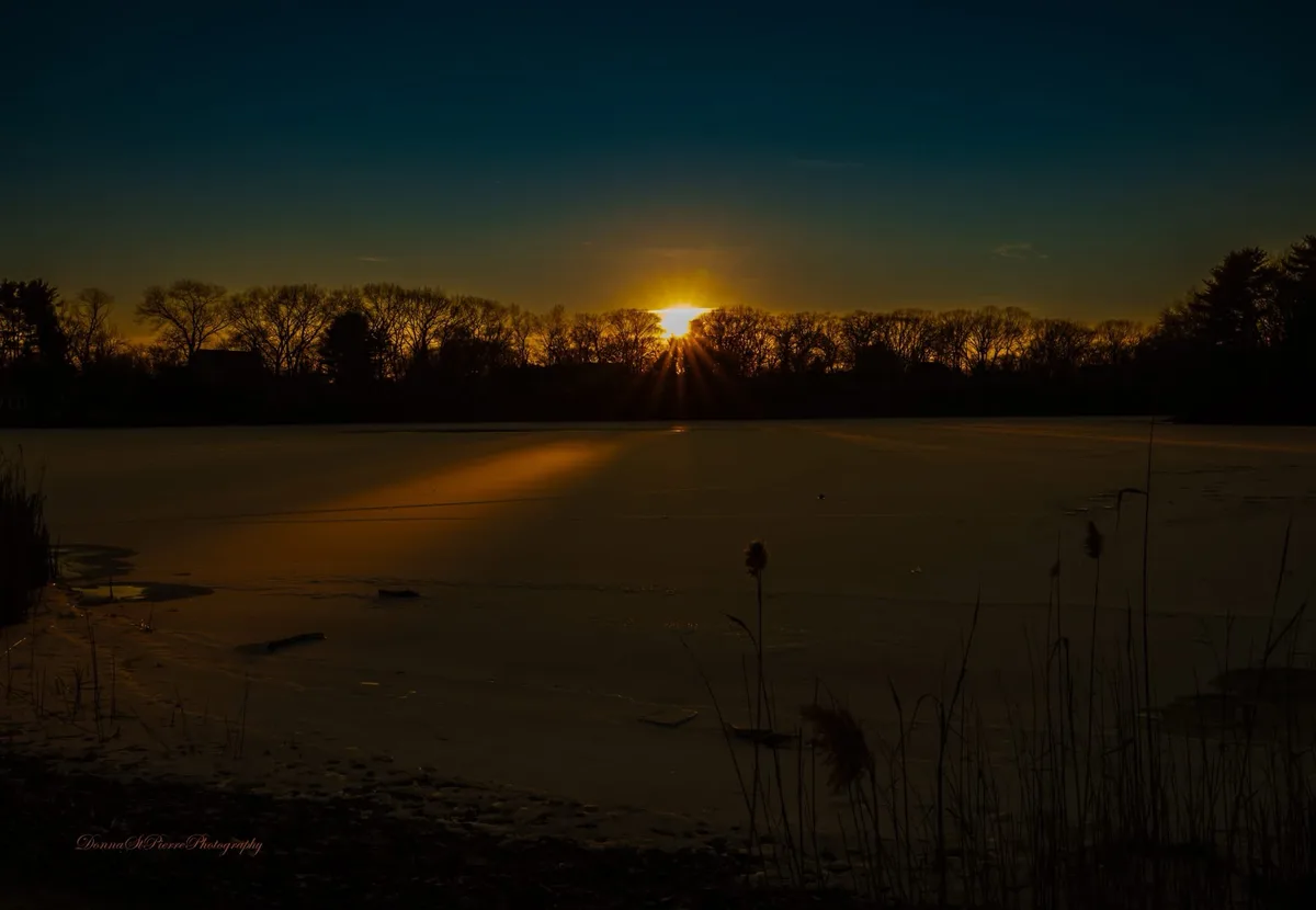 A serene photograph of a sunrise over a frozen lake, with the sun casting golden rays through silhouetted trees on the…
