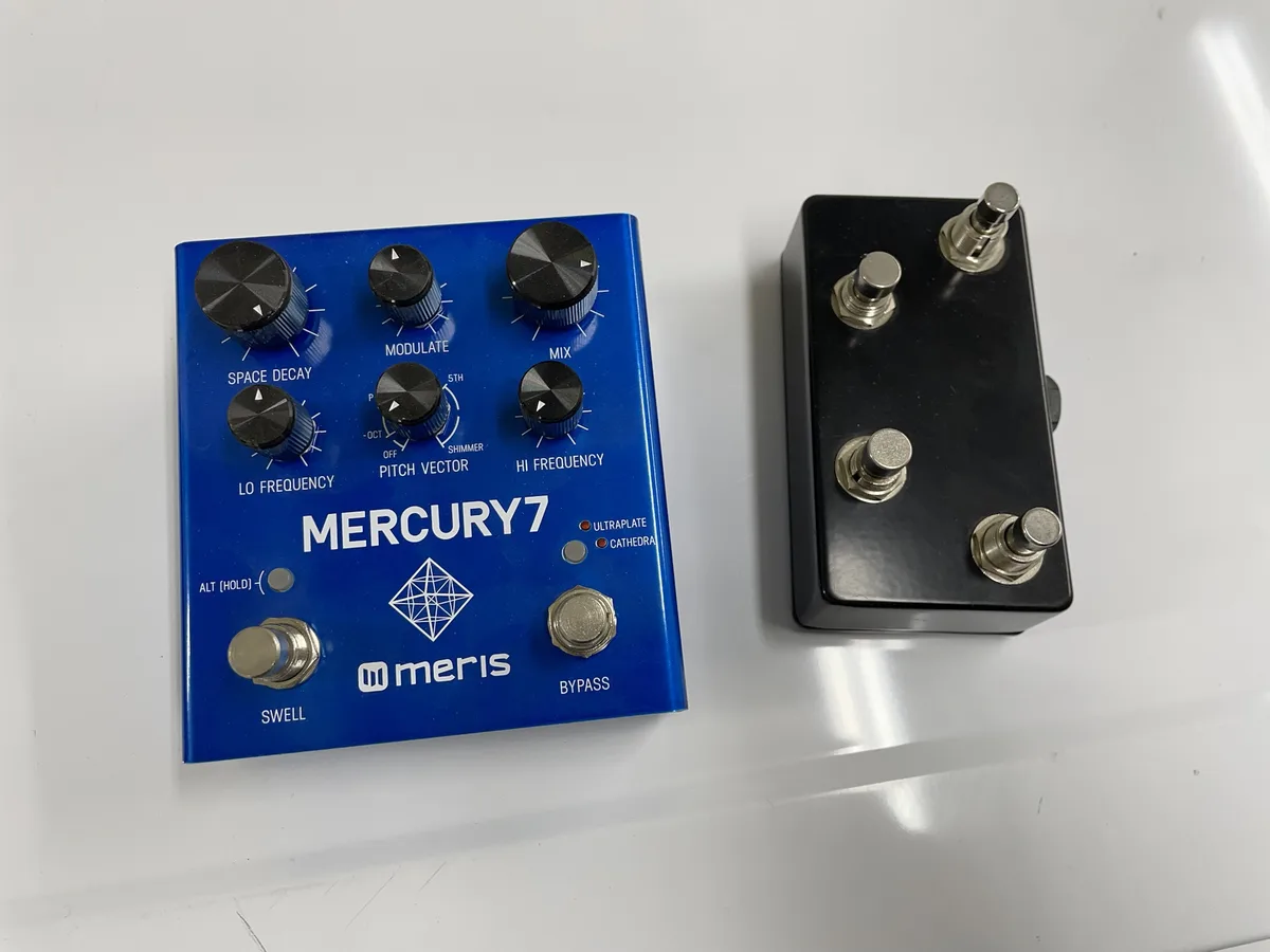 A blue Meris Mercury 7 guitar effects pedal with multiple knobs and footswitches sits next to a simple black pedal with four…