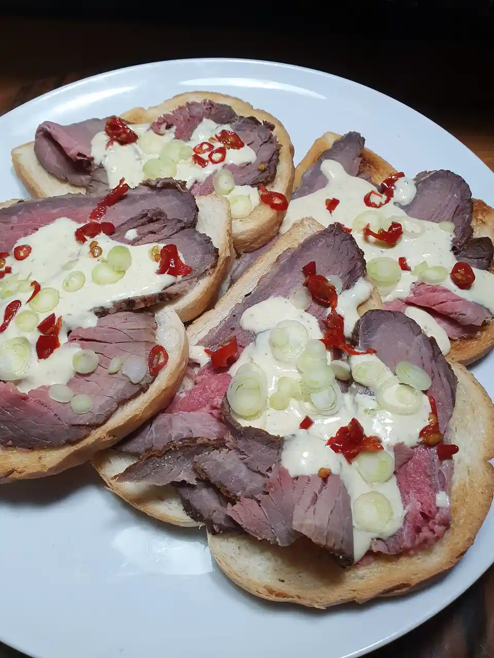 A white plate holds several open-faced sandwiches made with sliced roast beef, creamy white sauce, chopped green onions, and…