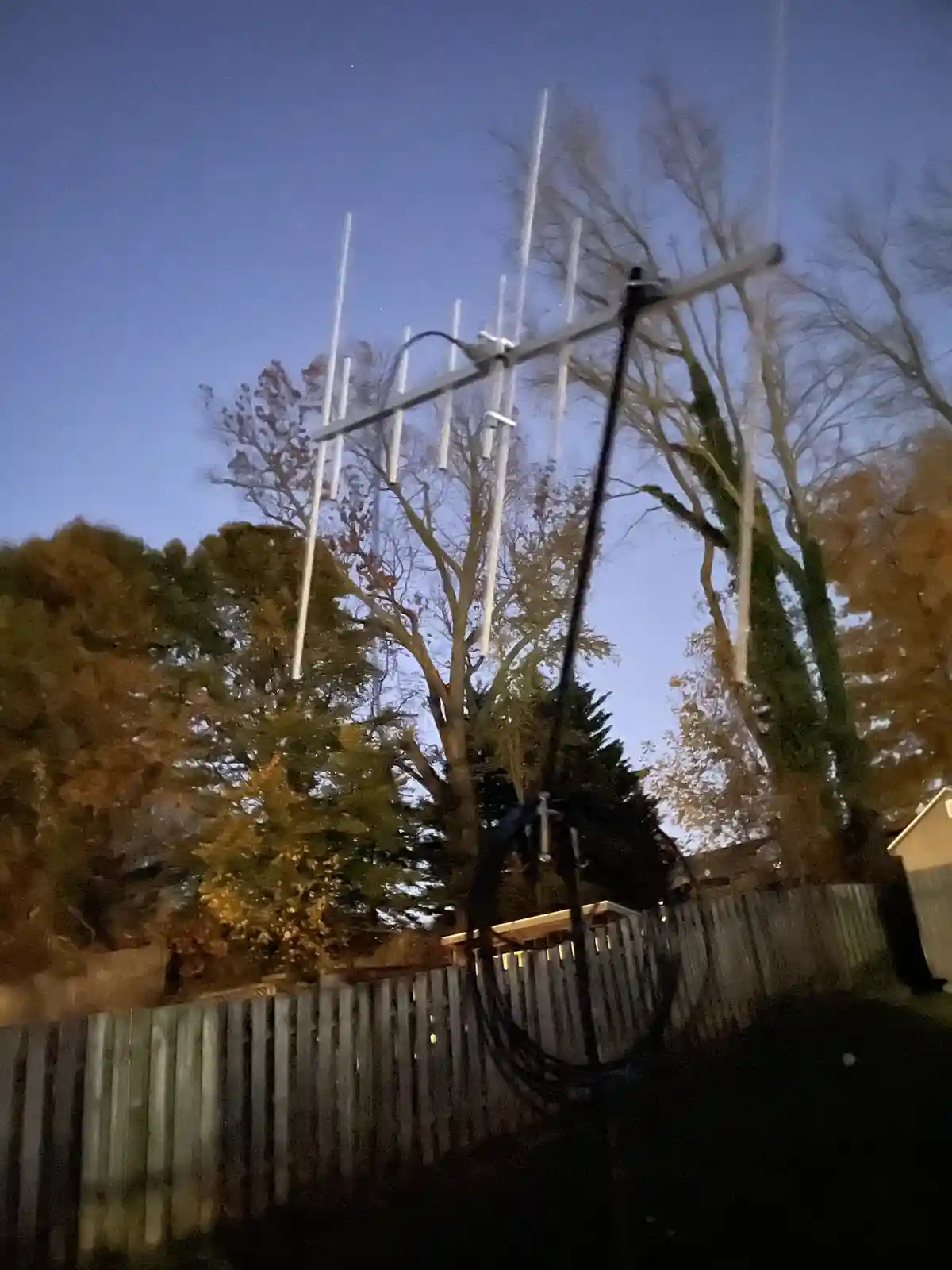 A blurry nighttime photo shows a white Yagi-Uda style TV antenna mounted on a pole in a backyard, with trees and a wooden…
