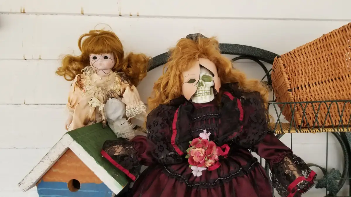 Two vintage dolls sit on a metal bench against a white wooden wall. The doll on the right wears a dark, ornate dress and…