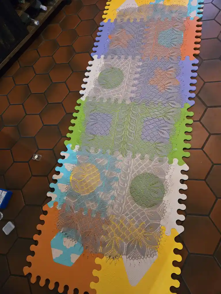 An overhead shot shows a large, intricate lace blanket laid out on a colorful, interlocking foam puzzle mat on a hexagonal…