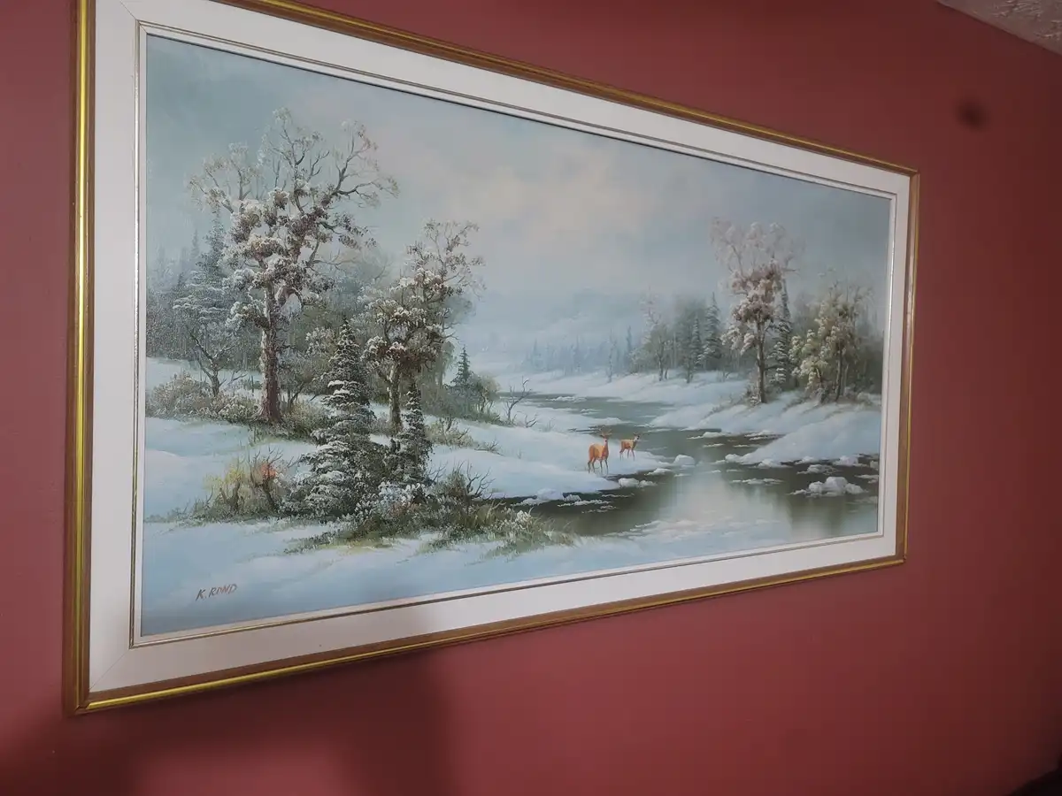 A framed painting of a snowy winter landscape hangs on a red wall, depicting two deer standing in a partially frozen river…