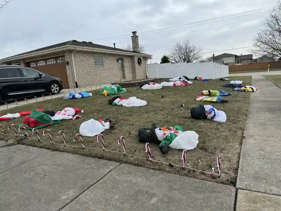 A front yard with deflated, crumpled Christmas decorations scattered across the grass, including Santa Claus, snowmen, and…