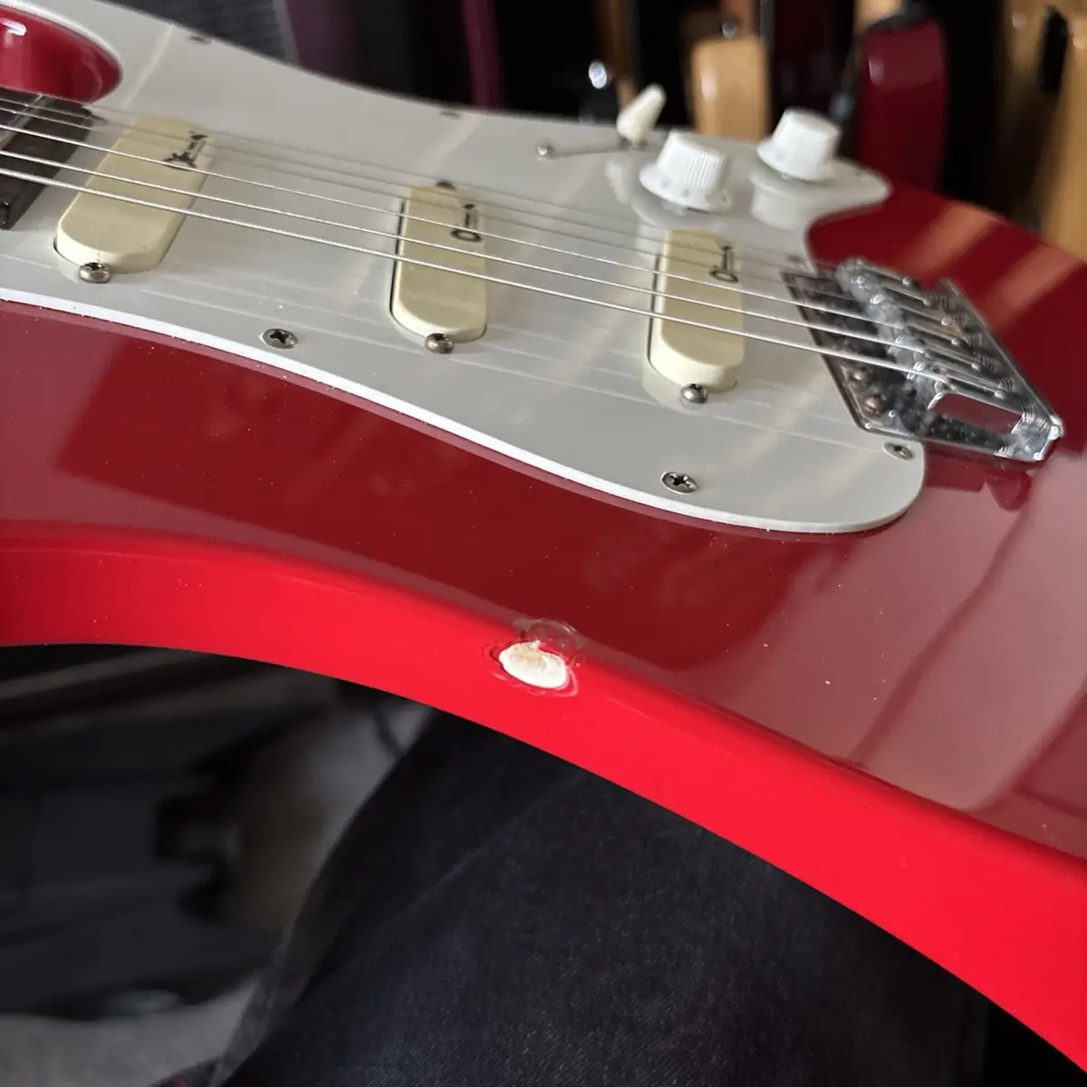 A close-up photo of a red electric guitar with a white pickguard, showing a small, circular hole punched through the body…