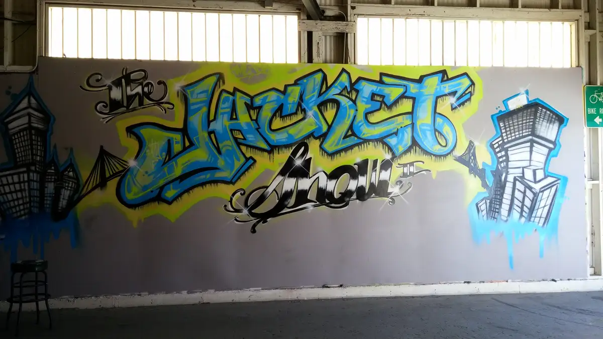 A large graffiti mural on a gray wall inside a warehouse-like space, featuring the words 'The Jackets Show' in stylized blue…