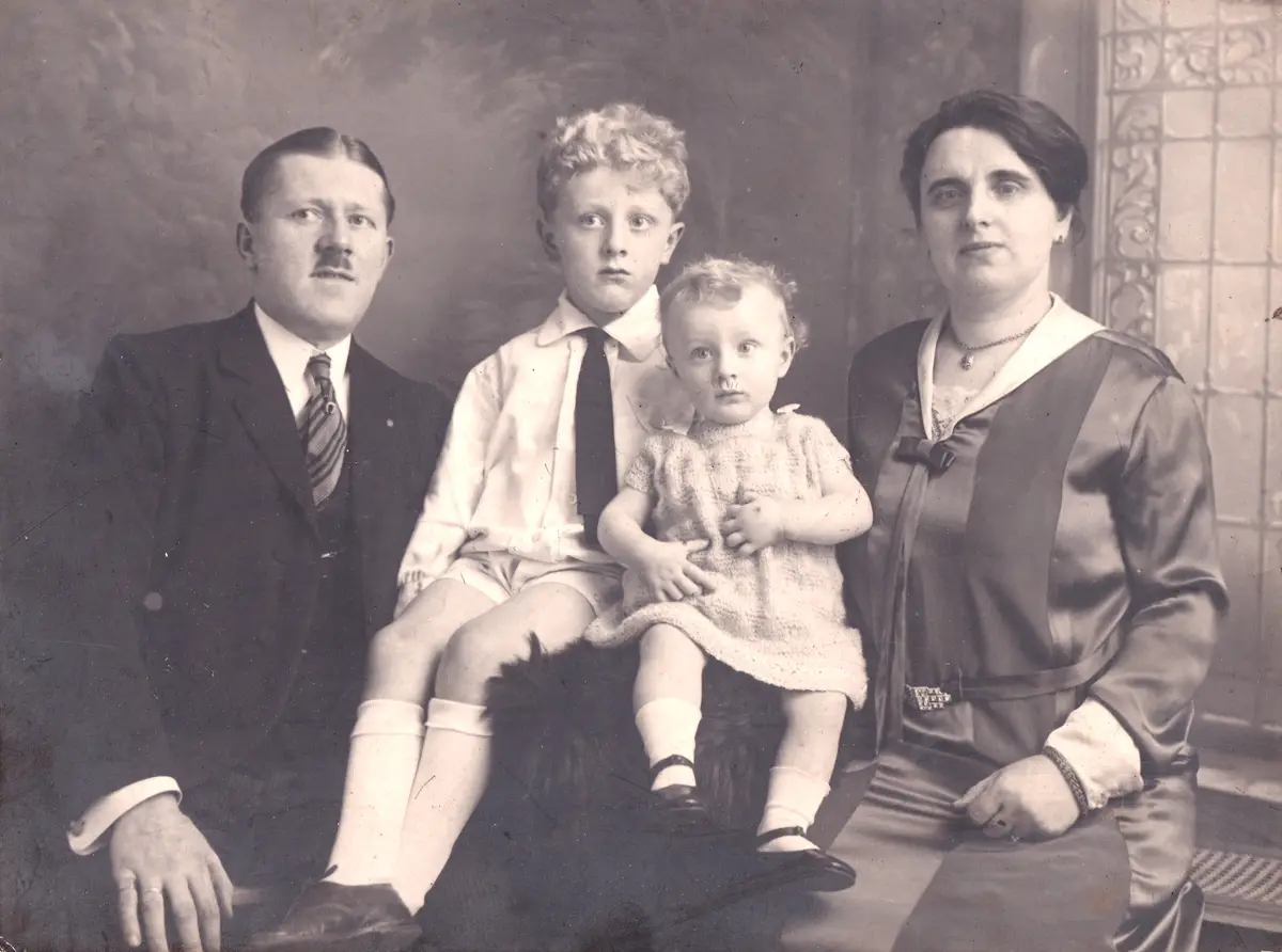 A vintage black-and-white family portrait shows a stern-looking man, woman, and two children. The toddler girl in the center…