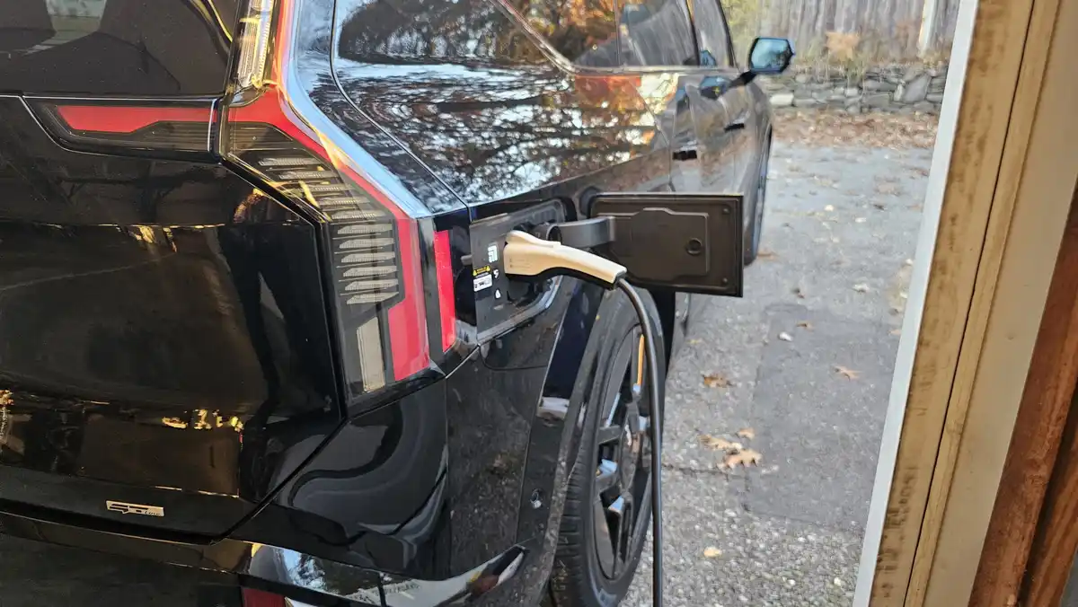 A black electric vehicle is plugged into a charging station on a driveway with fallen leaves, viewed from inside a house…