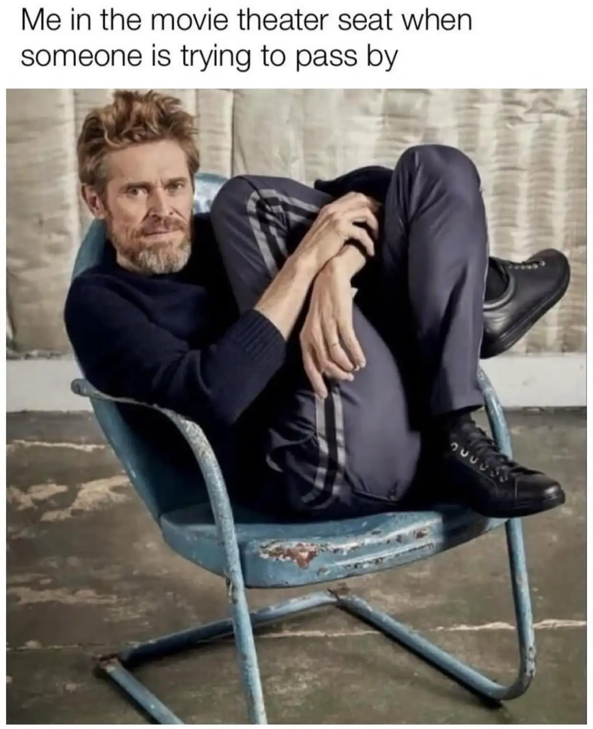 A man with a beard and tousled hair sits in a worn blue metal chair, legs crossed and arms wrapped around his knees, looking…