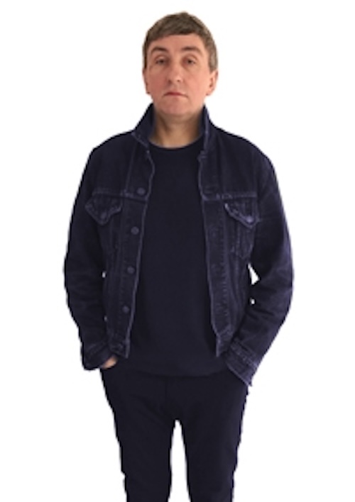 A man with short brown hair, wearing a dark denim jacket over a black t-shirt and black pants, stands against a white…