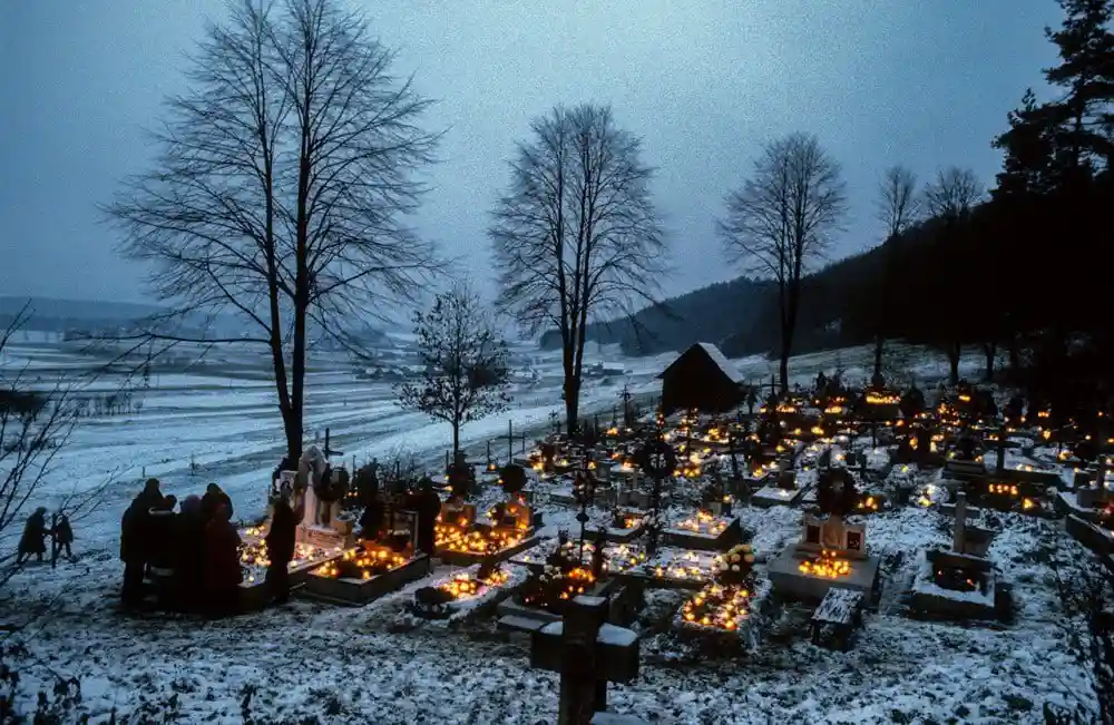 a snowy cemetery at dusk with many graves adorned with lit candles, silhouetted figures gathered near a central monument…