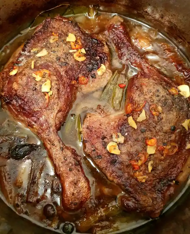 A close-up photo shows two cooked duck legs in a pot with a brown sauce, garnished with sliced red chilies and garlic. The…