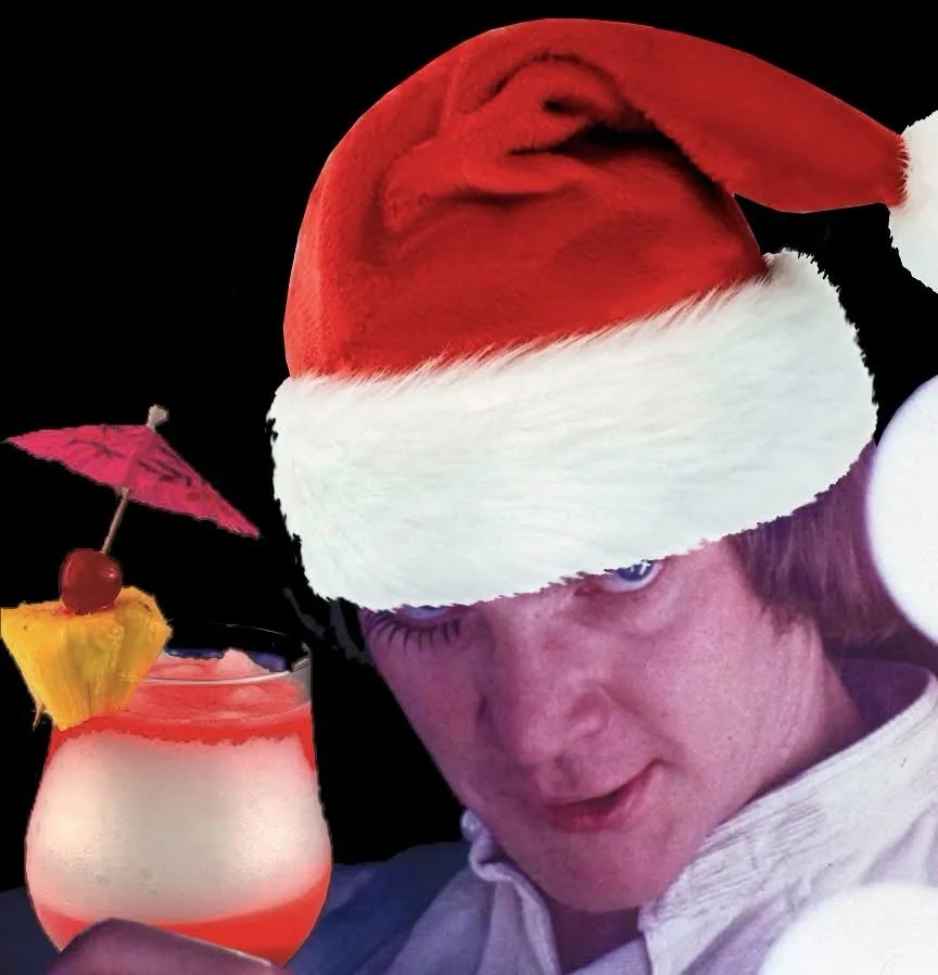 A man wearing a red and white Santa hat looks intently at a tropical cocktail with a pineapple wedge, cherry, and pink…