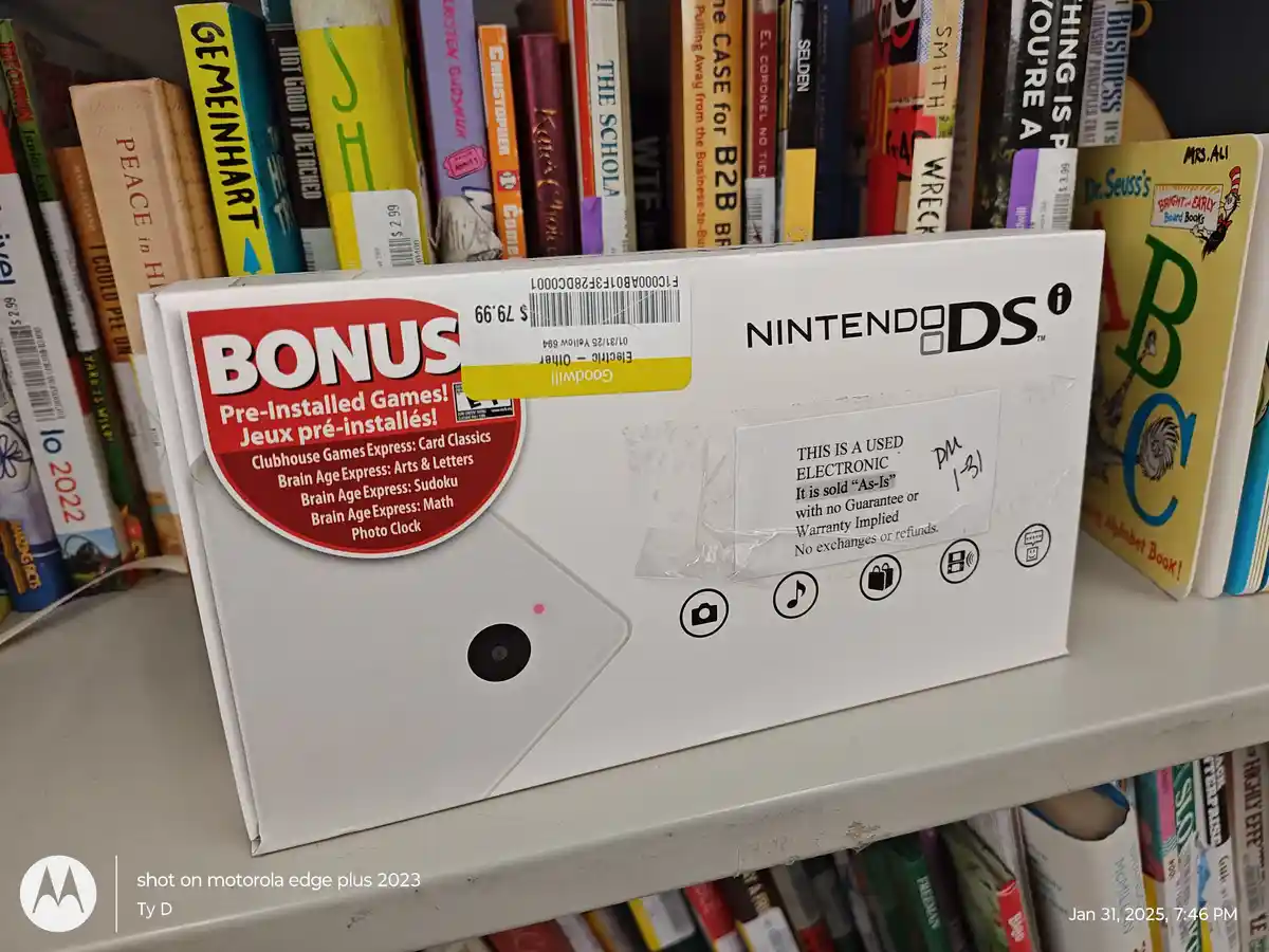A white Nintendo DS box sits on a shelf surrounded by books, with a 'BONUS Pre-Installed Games!' sticker and a 'Goodwill'…