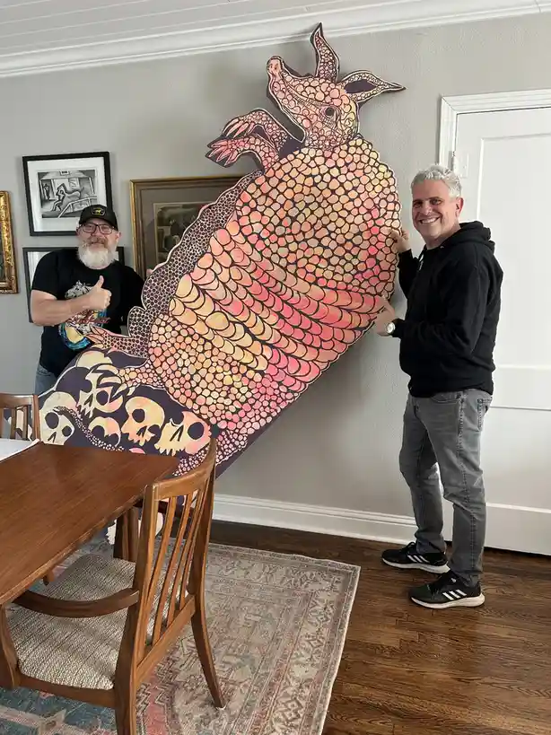 two men stand indoors holding up a large, stylized cutout of an armadillo with a gradient orange-pink shell and skulls at…