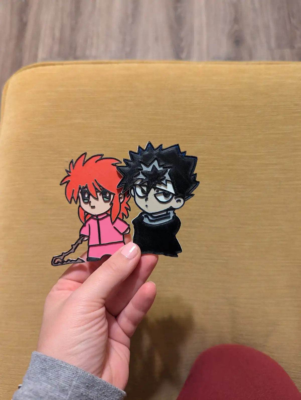 A hand holds two chibi-style anime character pins against a mustard-yellow fabric background. The pin on the left depicts a…