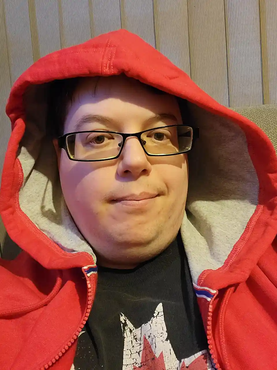 A person wearing a red hooded sweatshirt with the hood pulled up over their head, looking directly at the camera with a…
