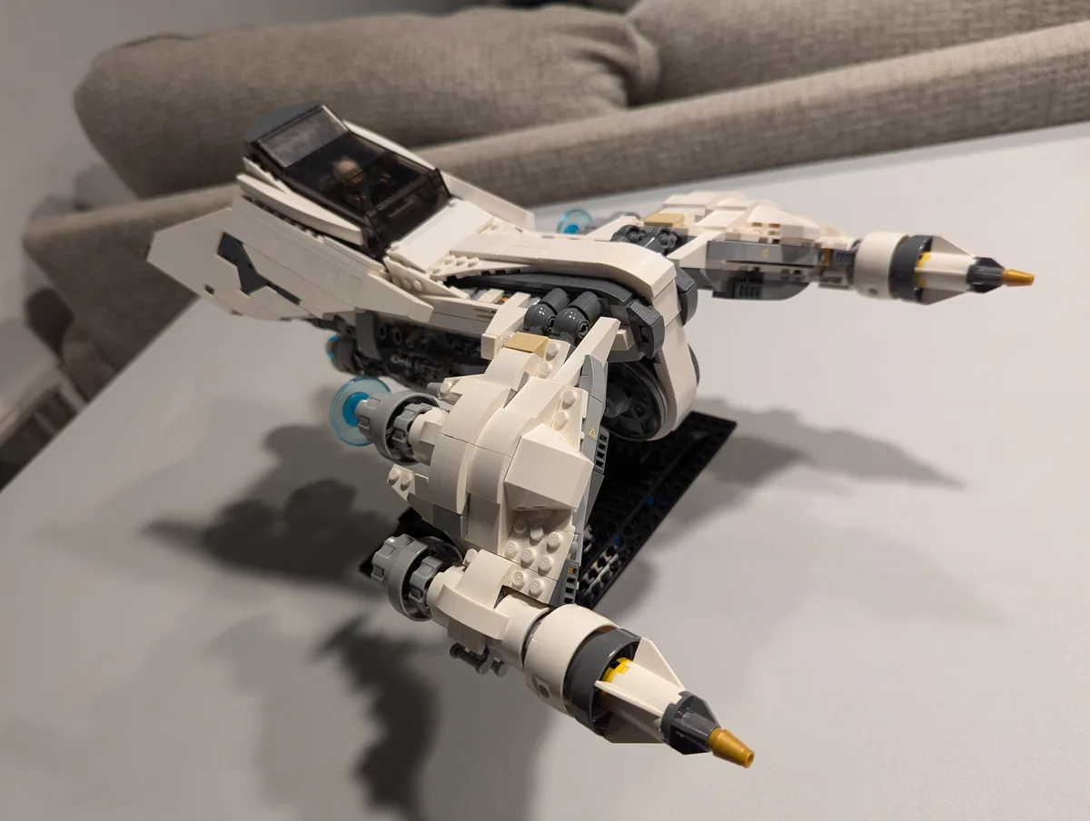 A detailed white, grey, and black LEGO Star Wars starfighter model is displayed on a white table, with a grey couch in the…
