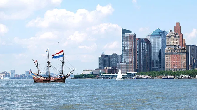 A historic Dutch sailing ship with a red, white, and blue flag sails on a body of water in front of a modern city skyline…