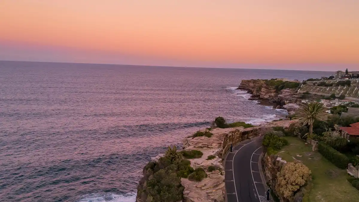 Vibrant sunrise hues splinter the horizon, illuminating the winding coastal road below. Towering cliffs plunge into crystal…