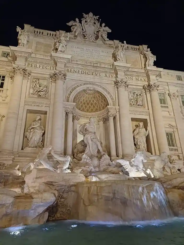 A nighttime photograph of the Trevi Fountain in Rome, Italy, illuminated by artificial lights against a dark sky. The…