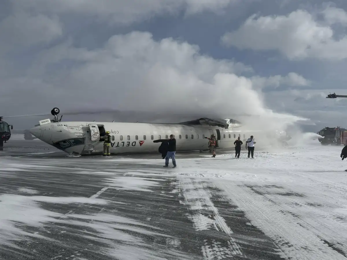 A Delta airplane is on fire on a snowy runway, with firefighters spraying water and people standing nearby. One man in the…