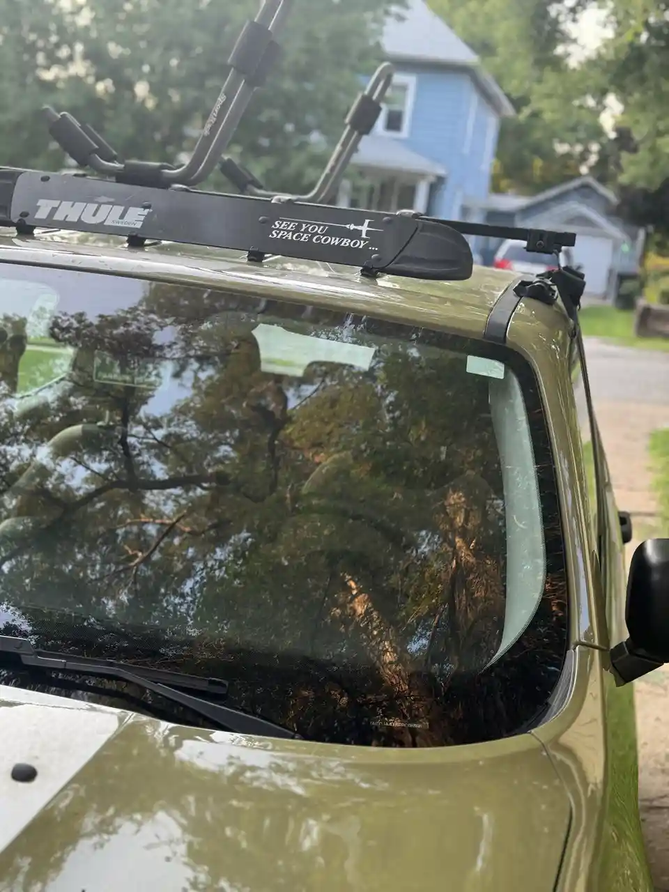 A close-up of the roof of an olive-green vehicle, showing a black Thule bike rack with a sticker that reads 'SEE YOU SPACE…