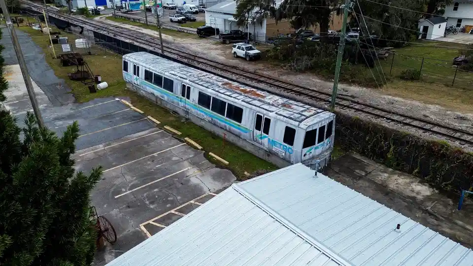 An aerial photo shows a weathered, rusted, and abandoned Metro train car lying horizontally on its side in a parking lot…