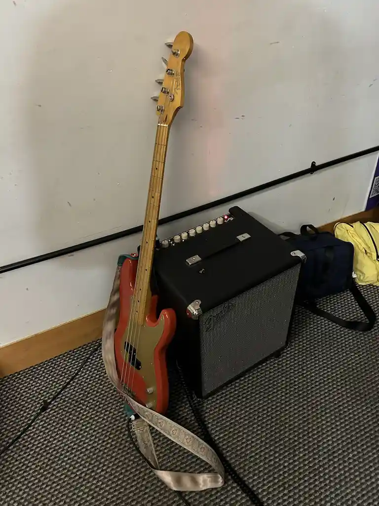 A red and gold Fender bass guitar leans against a black Fender amplifier on a grey carpeted floor, with a patterned strap…