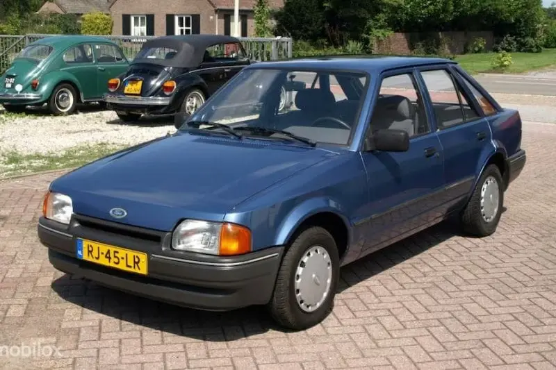 A blue 1980s Ford Escort sedan is parked on a brick driveway, with two vintage Volkswagen Beetles visible in the background…