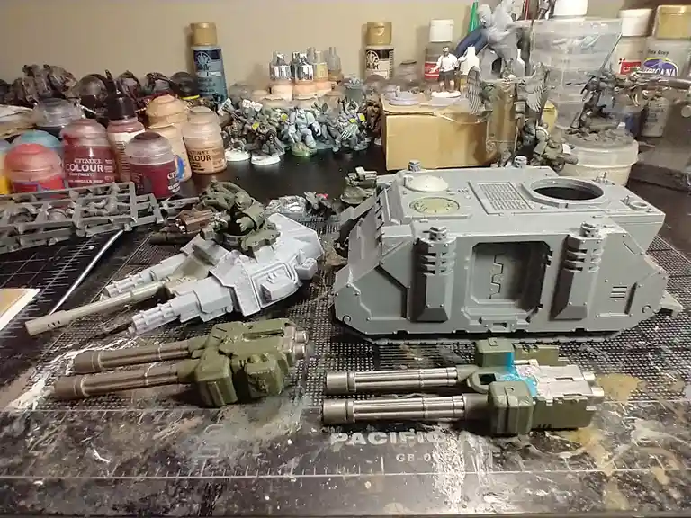 A cluttered hobbyist workspace shows unpainted and partially painted miniature war vehicles and weapons on a cutting mat…