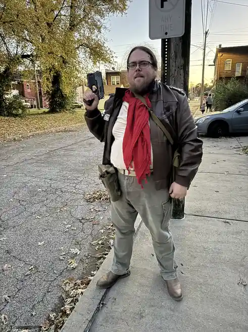 A man in a brown leather jacket, red scarf, and glasses stands on a sidewalk holding a black toy gun, posing next to a 'No…