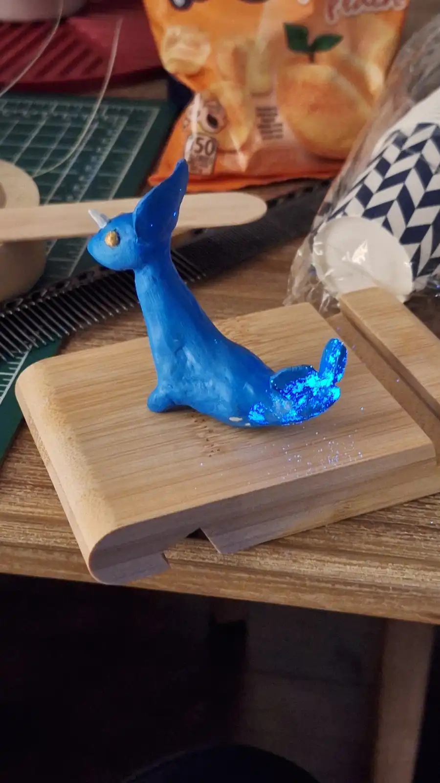 A handmade blue clay figurine of a small, stylized creature with a unicorn horn and glittery tail sits on a wooden stand…