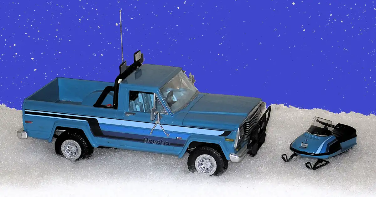 A blue toy pickup truck with 'Honcho' written on its side is parked in snow next to a matching blue toy snowmobile, both set…