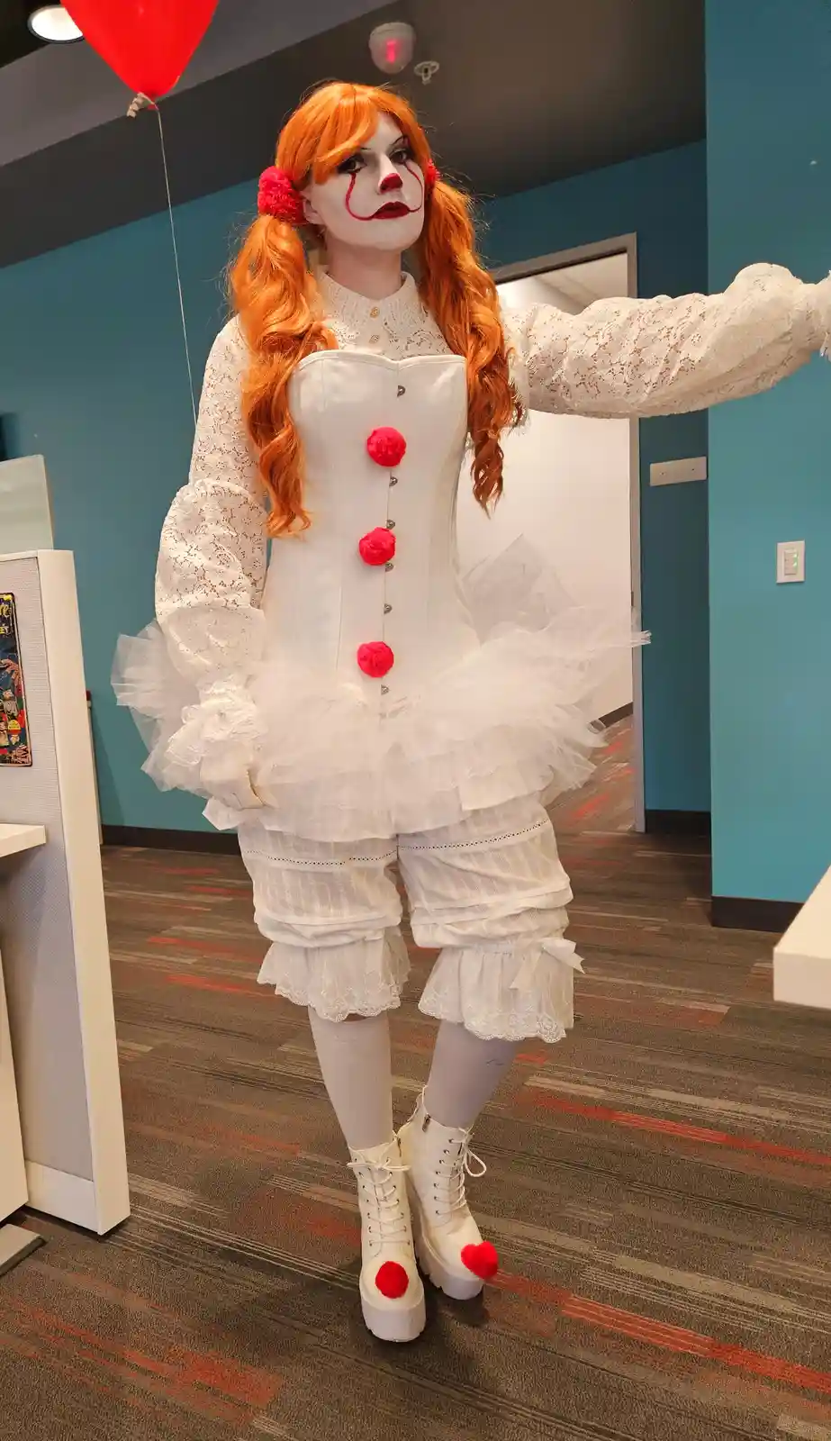 A person in a full clown costume with orange pigtails, white lace outfit, red pom-pom buttons, and red nose makeup stands…