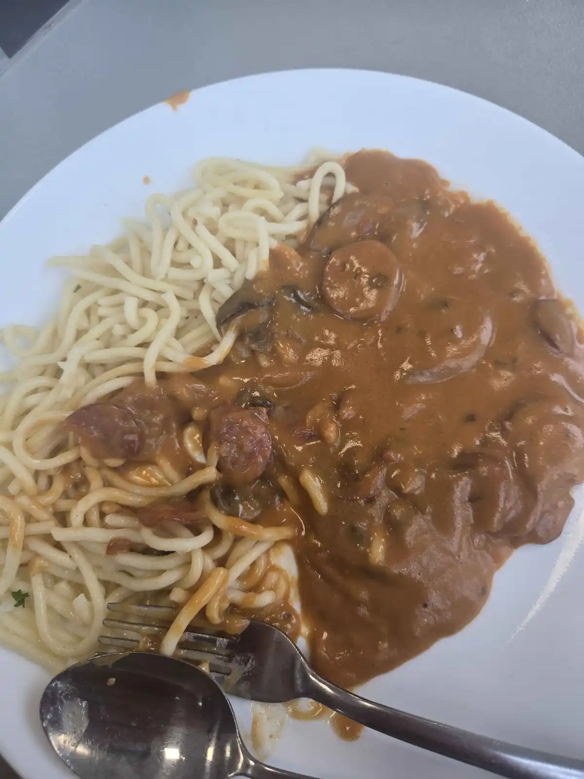 A white plate holds noodles partially covered in a thick brown gravy with chunks of meat and mushrooms, with a fork and…