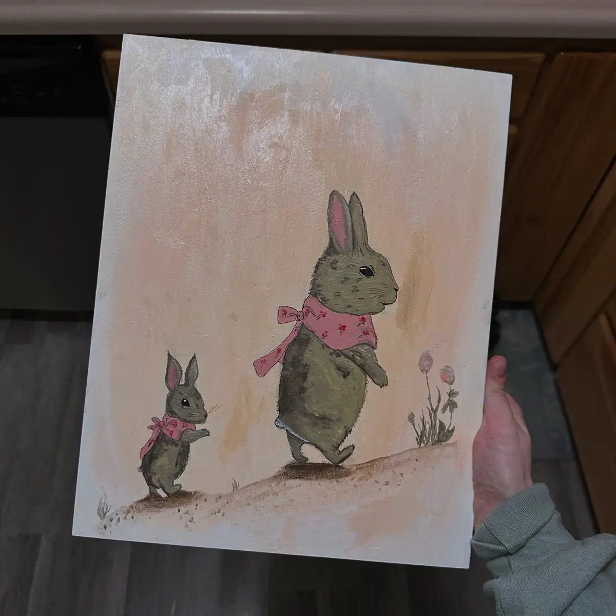A person holds up a painting depicting two rabbits, one large and one small, both wearing pink floral scarves, standing on a…