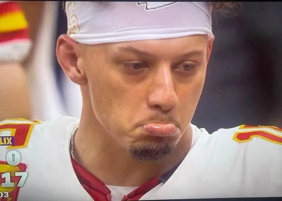 A close-up of a football player wearing a white jersey with red and yellow accents and a white headband, looking down with a…