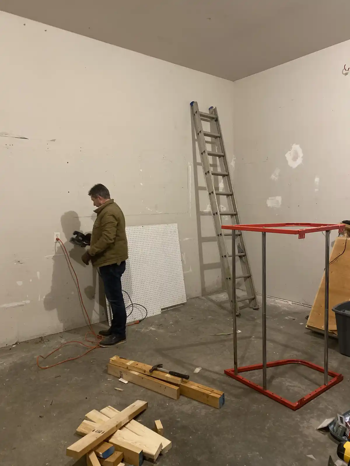 A man in a brown jacket and jeans stands in a room with unfinished walls, holding a power tool plugged into an outlet…