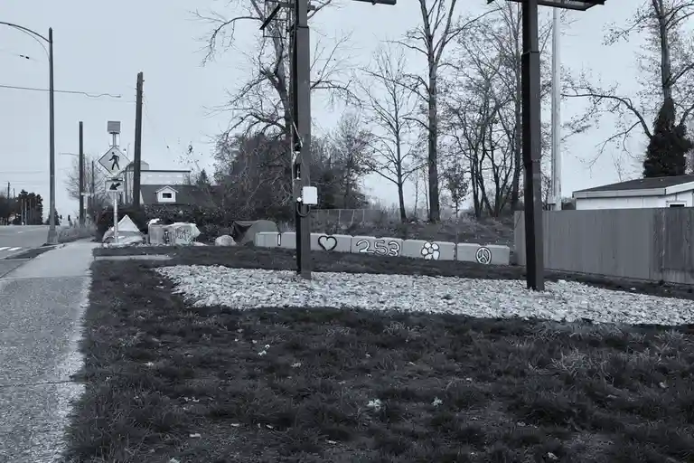 A black-and-white photo of a roadside area with a concrete barrier painted with a heart, the number 253, a flower, and a…