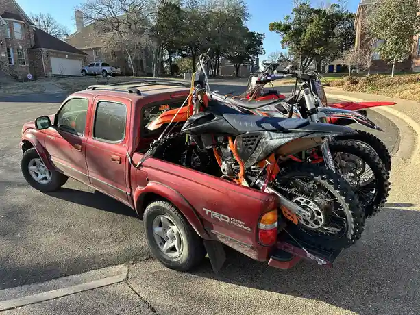 A red Toyota Tacoma pickup truck is parked on a residential street, overloaded with at least three dirt bikes strapped into…