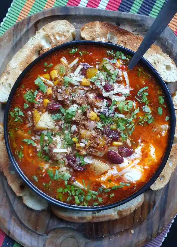 A top-down photo of a black bowl filled with a thick, red stew containing ground meat, kidney beans, corn, potatoes…
